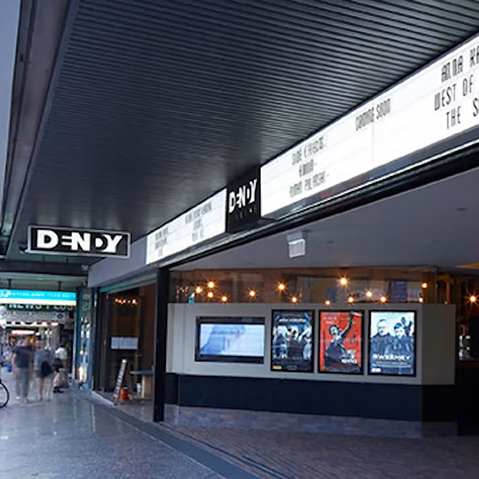 The streetfront of the Dendy Newtown cinema featuring the sign on the ceiling above, listed current movie titles and movie posters on the wall opposite
